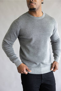 A man wearing a light gray ribbed knit crewneck sweater and black pants, standing against a neutral gray background.