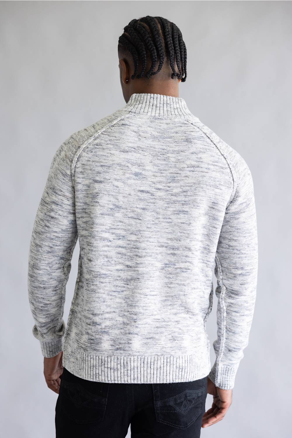 A man viewed from behind wearing a light gray and white heathered knit sweater with a mock neck and raglan sleeves, paired with black pants, against a neutral gray background.