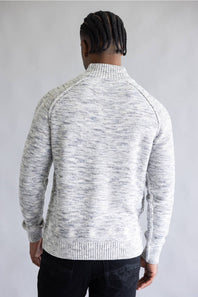 A man viewed from behind wearing a light gray and white heathered knit sweater with a mock neck and raglan sleeves, paired with black pants, against a neutral gray background.