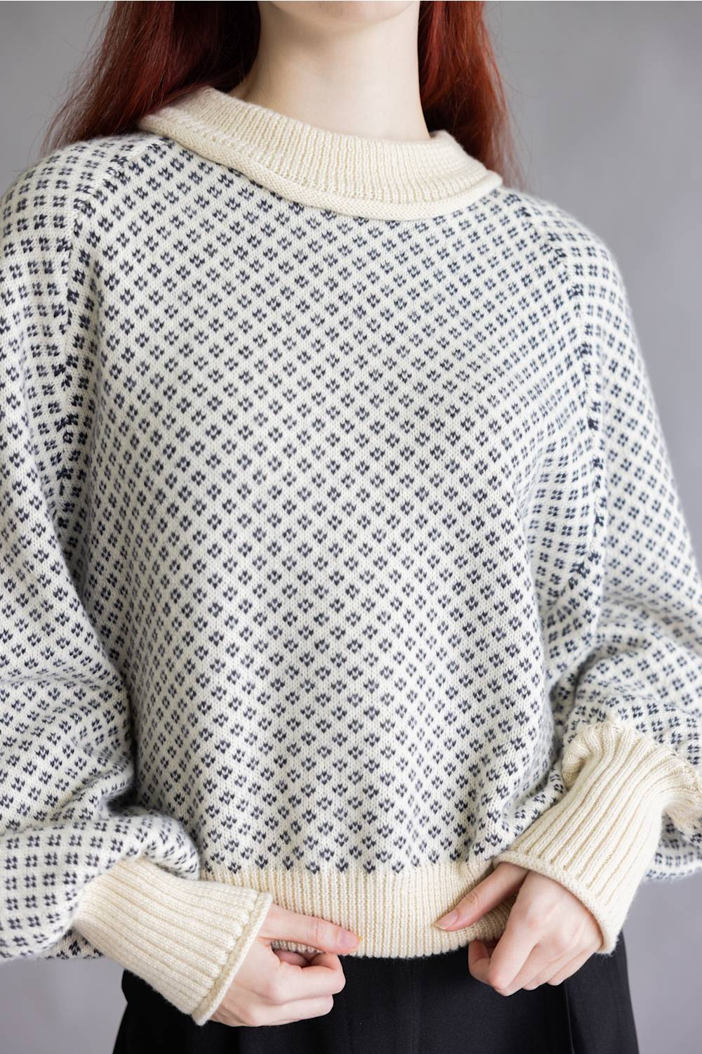 A close-up of a person wearing a cream-colored knit sweater with a small repeating dark geometric pattern. The sweater features a ribbed crew neck, cuffs, and hem, and is shown against a neutral gray background.