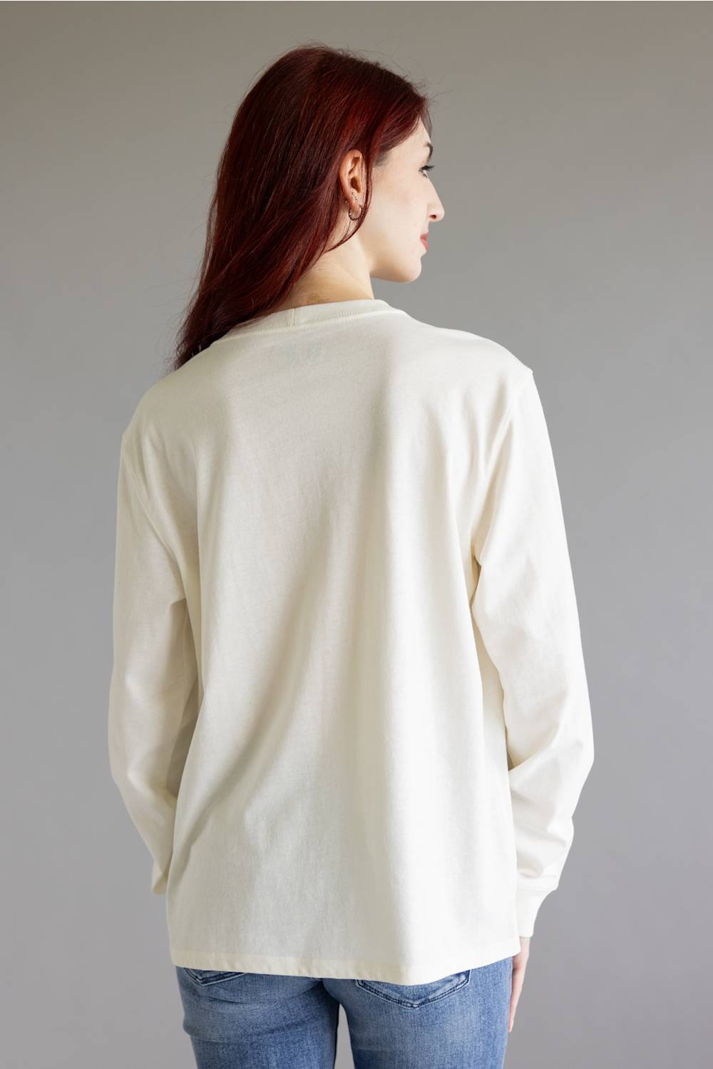 A woman with long red hair is shown from the back wearing a cream-colored long-sleeve top and blue jeans against a plain gray studio background.