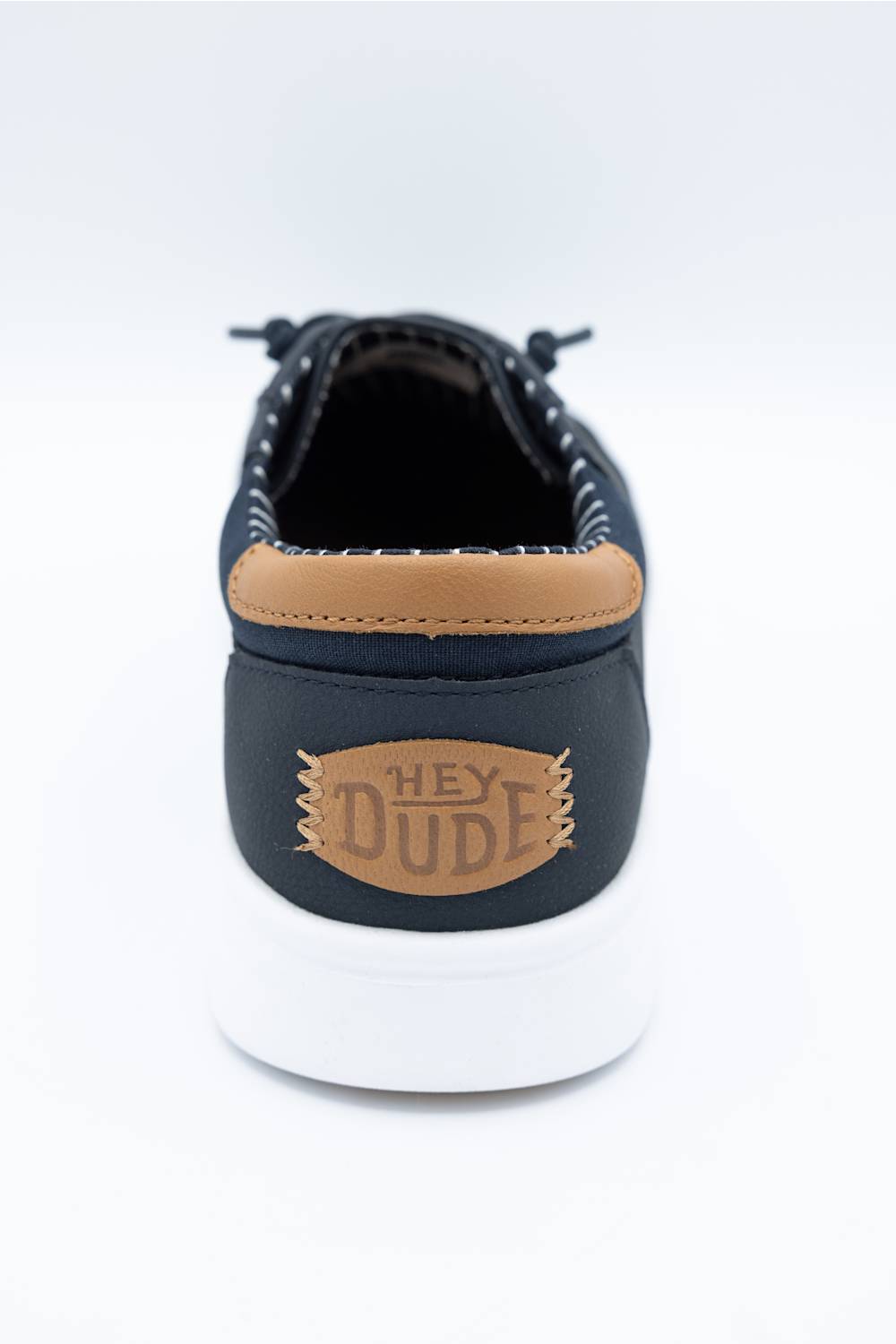 A single low-top sneaker shown from the back, featuring a black upper, a tan leather heel tab with 'HEY DUDE' branding, and a thick white rubber sole, photographed on a clean white background.