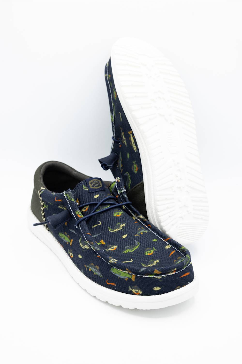 A pair of low-top casual shoes in navy blue canvas with a green, orange, and yellow fish pattern, featuring a white sole. One shoe is shown from the side displaying the pattern, while the other is positioned behind it showing the white outsole.