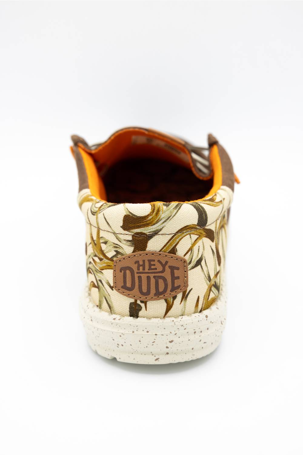A single low-top sneaker shown from the back on a white background, featuring a cream and brown abstract patterned canvas upper, a brown leather patch with 'HEY DUDE' text, and a speckled off-white rubber sole.