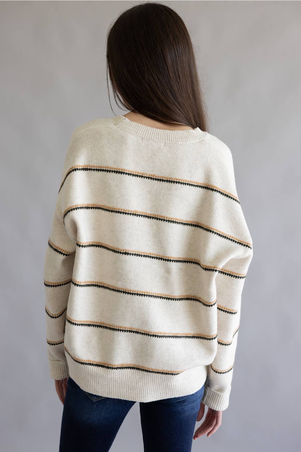A woman viewed from behind wearing a cream-colored knit sweater with thin horizontal stripes in black and tan, paired with dark blue jeans, against a plain gray studio background.