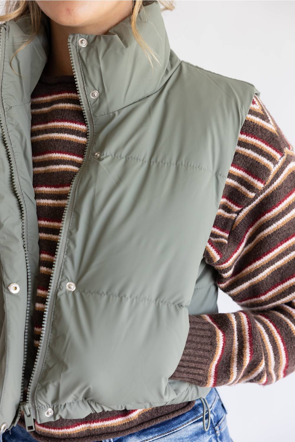 A close-up of a person wearing an olive green puffer vest over a brown, red, and cream striped knit sweater, paired with blue jeans.