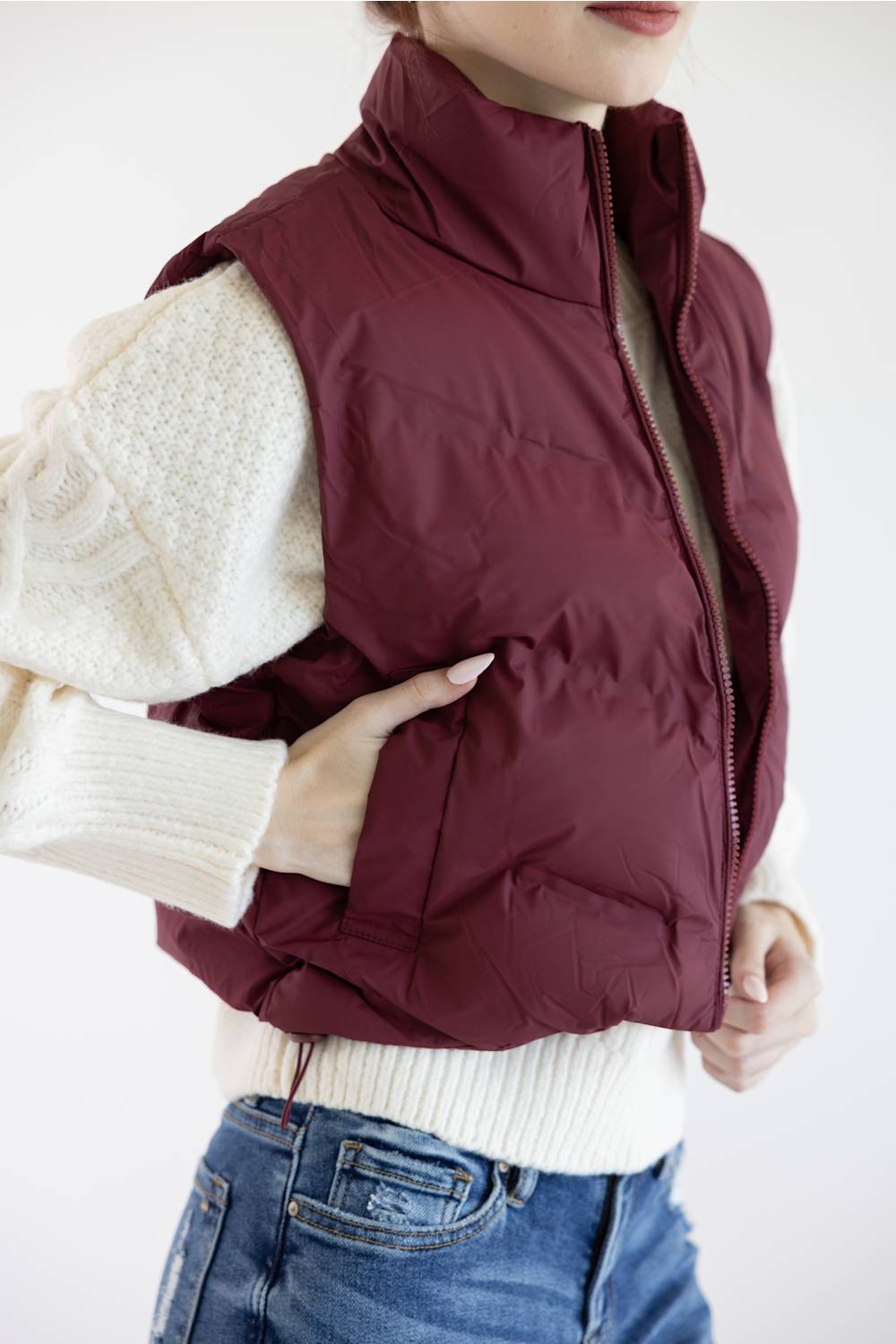 A woman wearing a sleeveless quilted puffer vest in deep burgundy over a cream knit sweater and blue jeans, shown from the chest down against a plain light background.