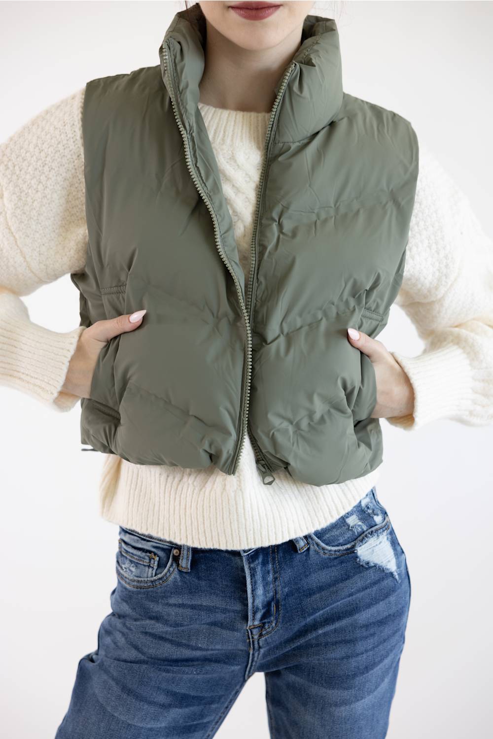 A person wearing an olive green puffer vest over a cream knit sweater and blue jeans, shown from the mouth to the upper thighs against a plain light background.