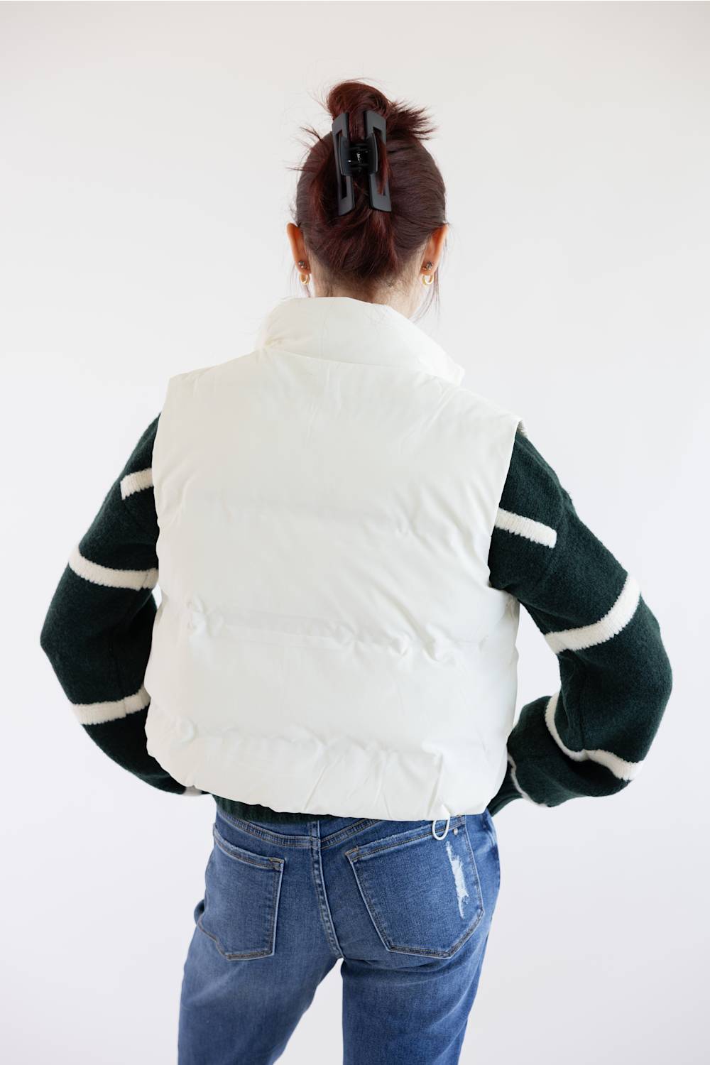A person shown from behind wearing a white puffer vest over a dark green and white striped sweater, paired with blue jeans, against a plain white background.