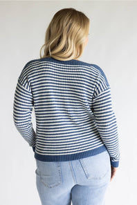 A woman viewed from behind wearing a navy and white striped knit sweater with light blue jeans, set against a plain light gray background.