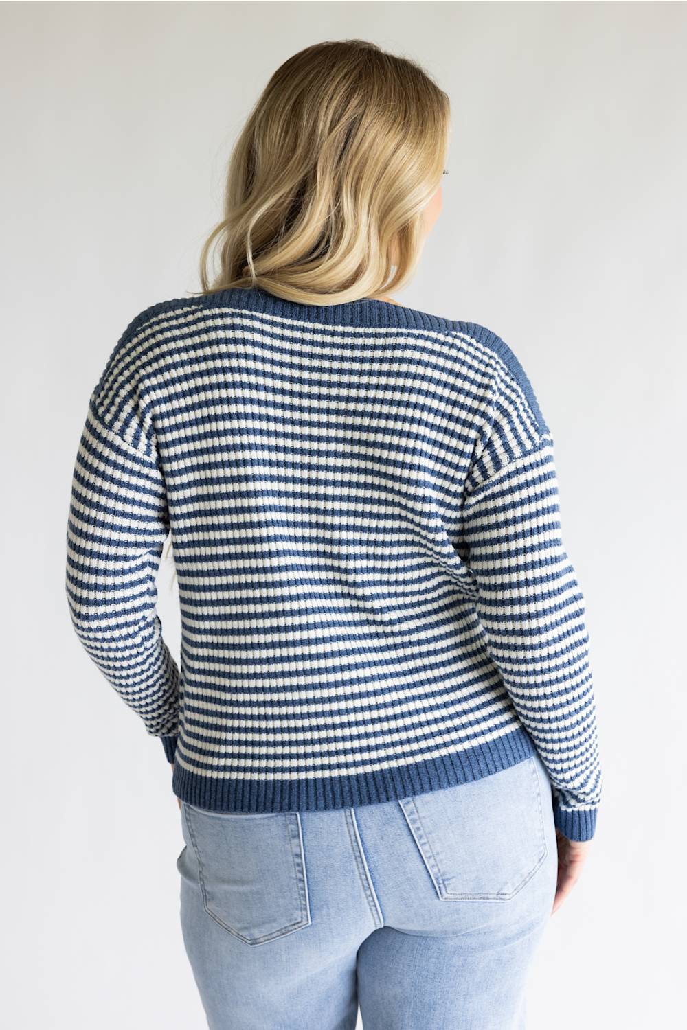 A woman viewed from behind wearing a navy and white striped knit sweater with light blue jeans, set against a plain light gray background.