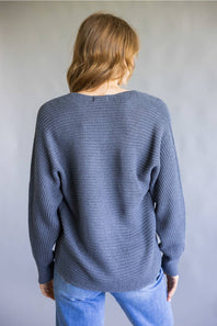 A woman viewed from behind wearing a slate blue ribbed knit sweater and light blue jeans, standing against a neutral gray background.
