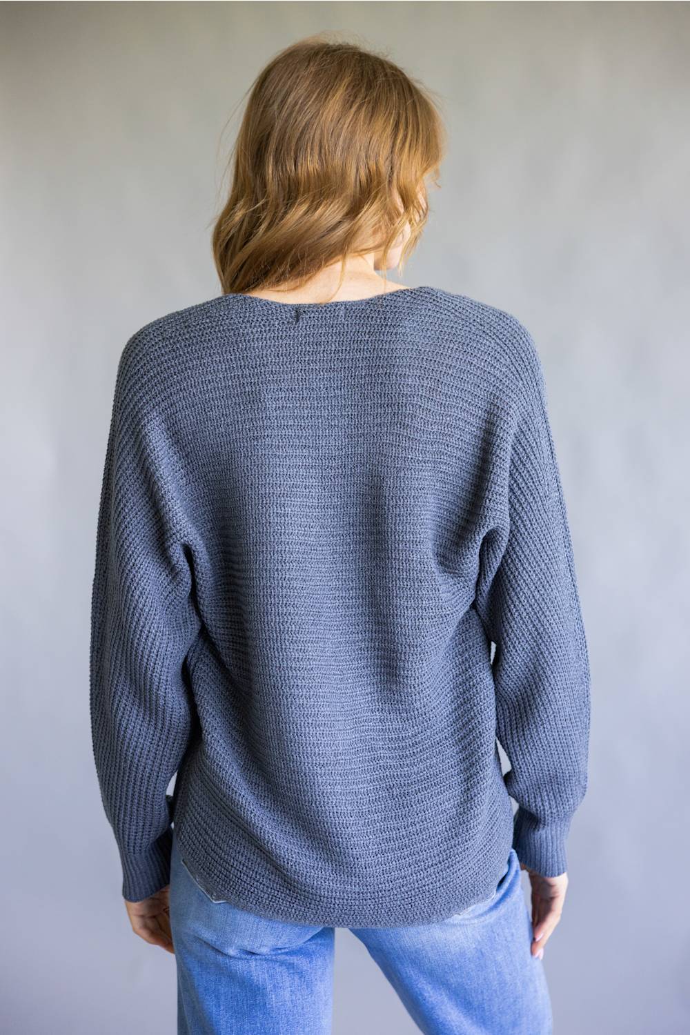 A woman viewed from behind wearing a slate blue ribbed knit sweater and light blue jeans, standing against a neutral gray background.