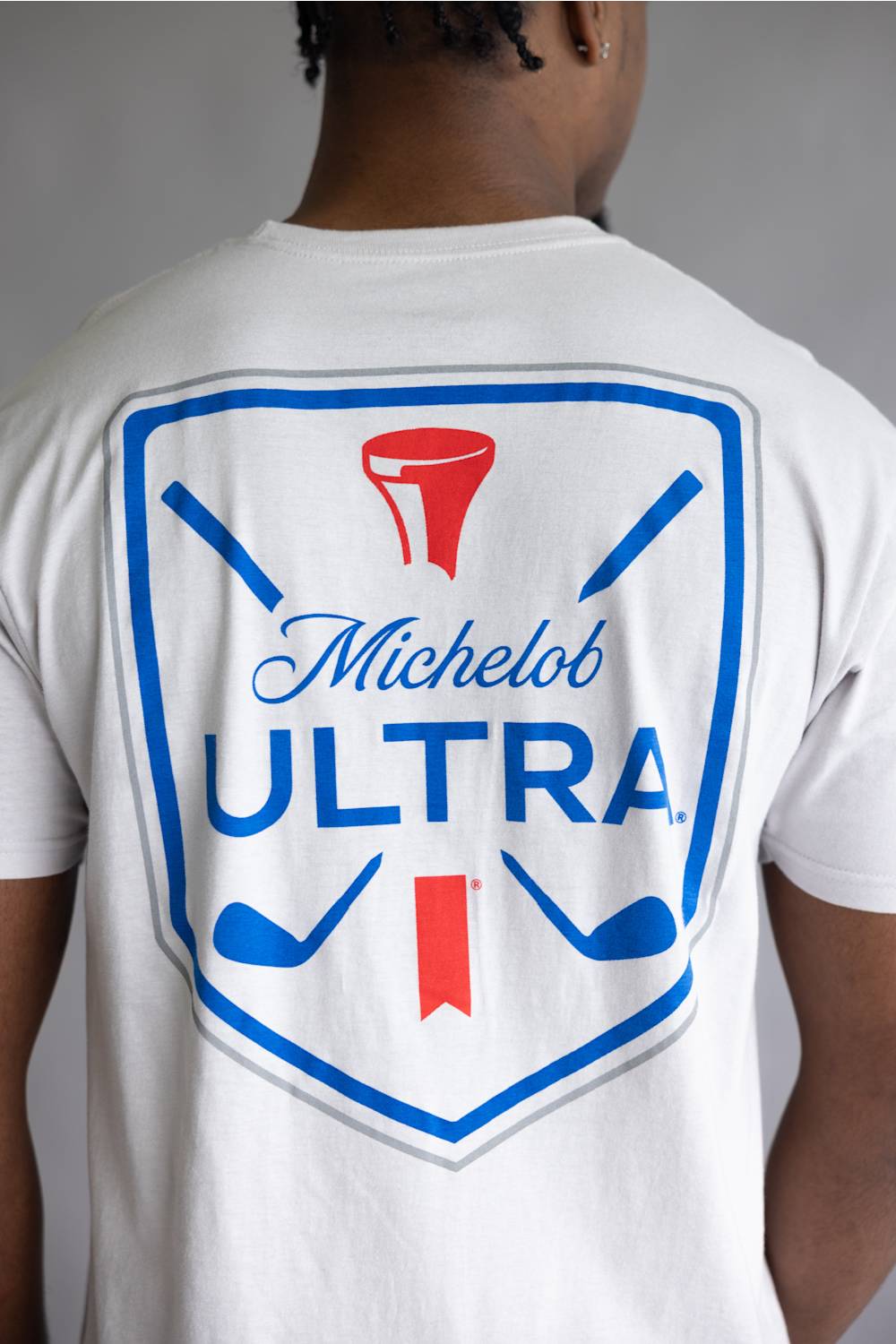 A person viewed from the back wearing a white short-sleeve T-shirt with a large blue and red Michelob ULTRA golf graphic printed on the back.