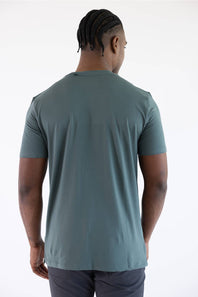 Back view of a man wearing the 7Diamonds Core Crew T-Shirt in Drake green, paired with grey pants against a white background.