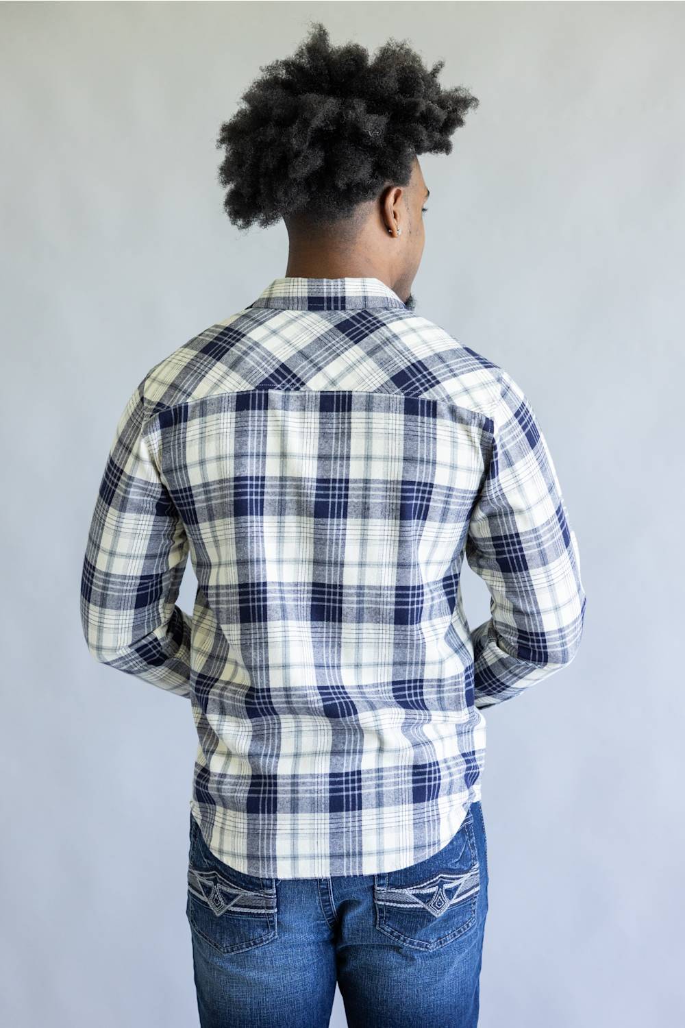 A man viewed from behind wearing a long-sleeve plaid button-up shirt in white, navy, and gray, paired with blue jeans, against a plain light gray background.