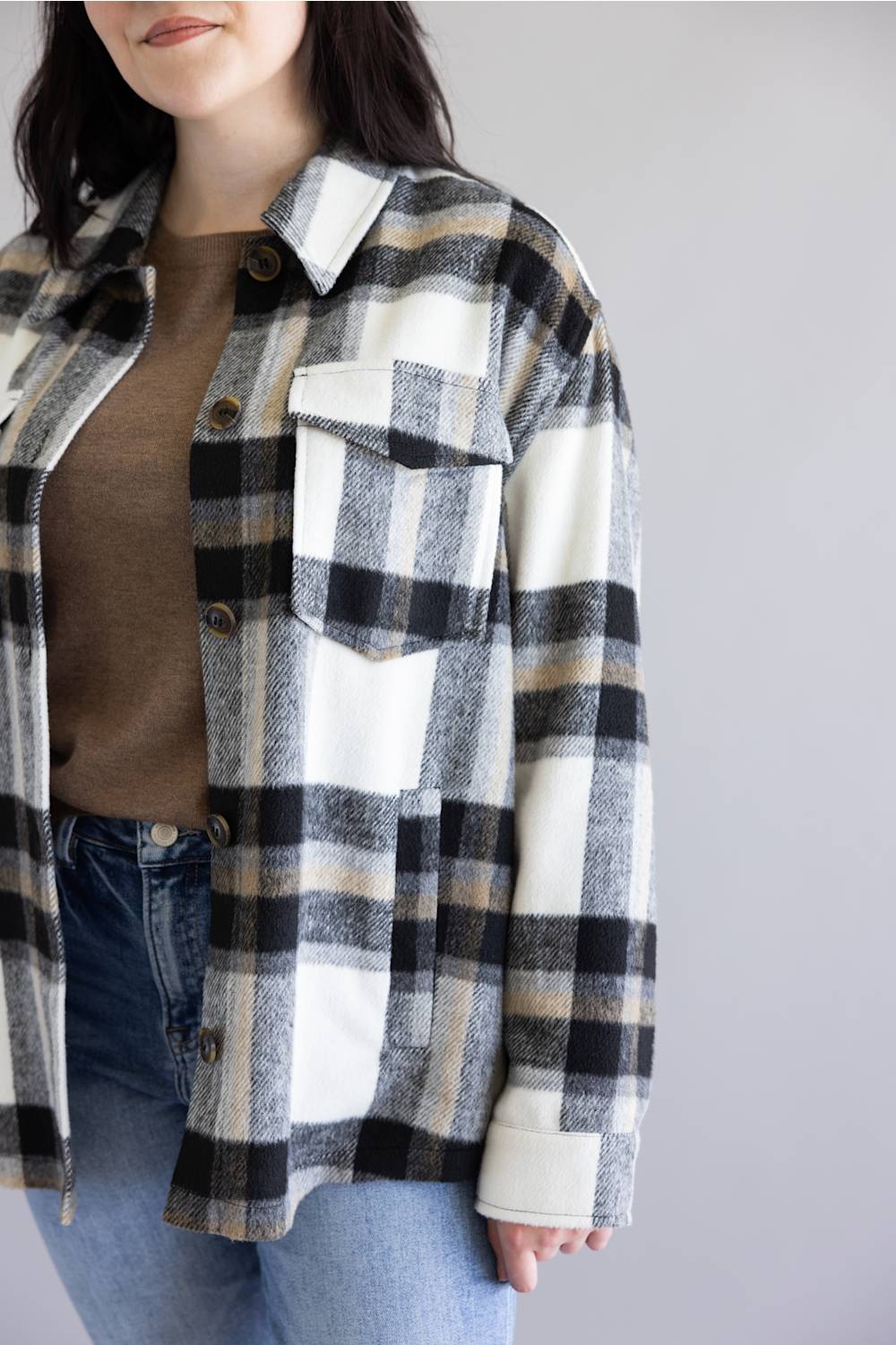 A woman wearing an oversized black, beige, and white plaid flannel shirt over a brown top and blue jeans, shown from the chin down against a plain light gray background.