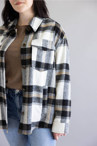 A woman wearing an oversized black, beige, and white plaid flannel shirt over a brown top and blue jeans, shown from the chin down against a plain light gray background.