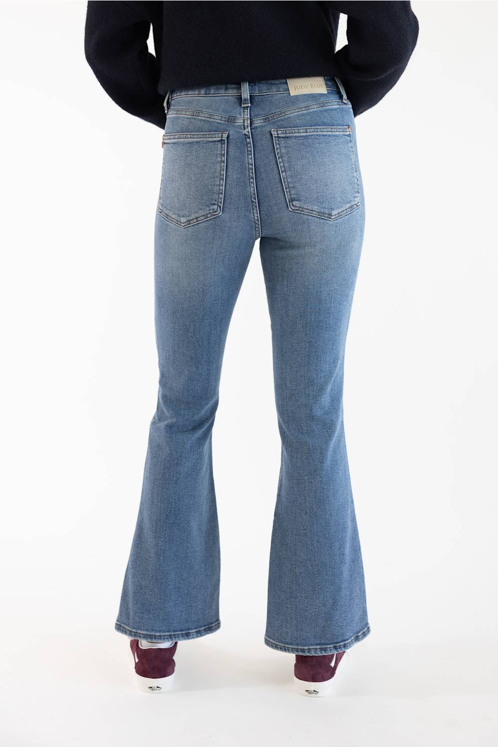 Rear view of a person wearing light-wash blue flare jeans with a black top and burgundy shoes, standing against a plain white studio background.