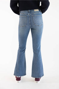 Rear view of a person wearing light-wash blue flare jeans with a black top and burgundy shoes, standing against a plain white studio background.