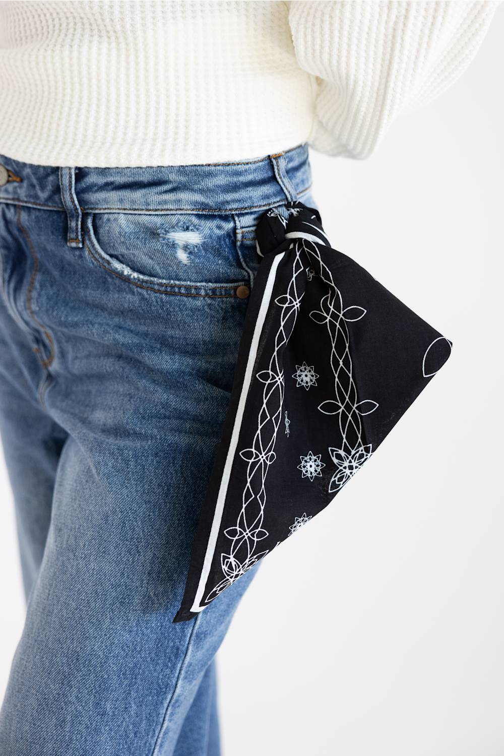 A black bandana with white floral and snowflake patterns tied around the waist of a person wearing blue jeans and a white ribbed sweater, shown against a plain white background.