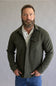 A man with a full beard wearing an olive green button-down shirt jacket over a cream button-down shirt and blue jeans, standing against a neutral gray studio background.