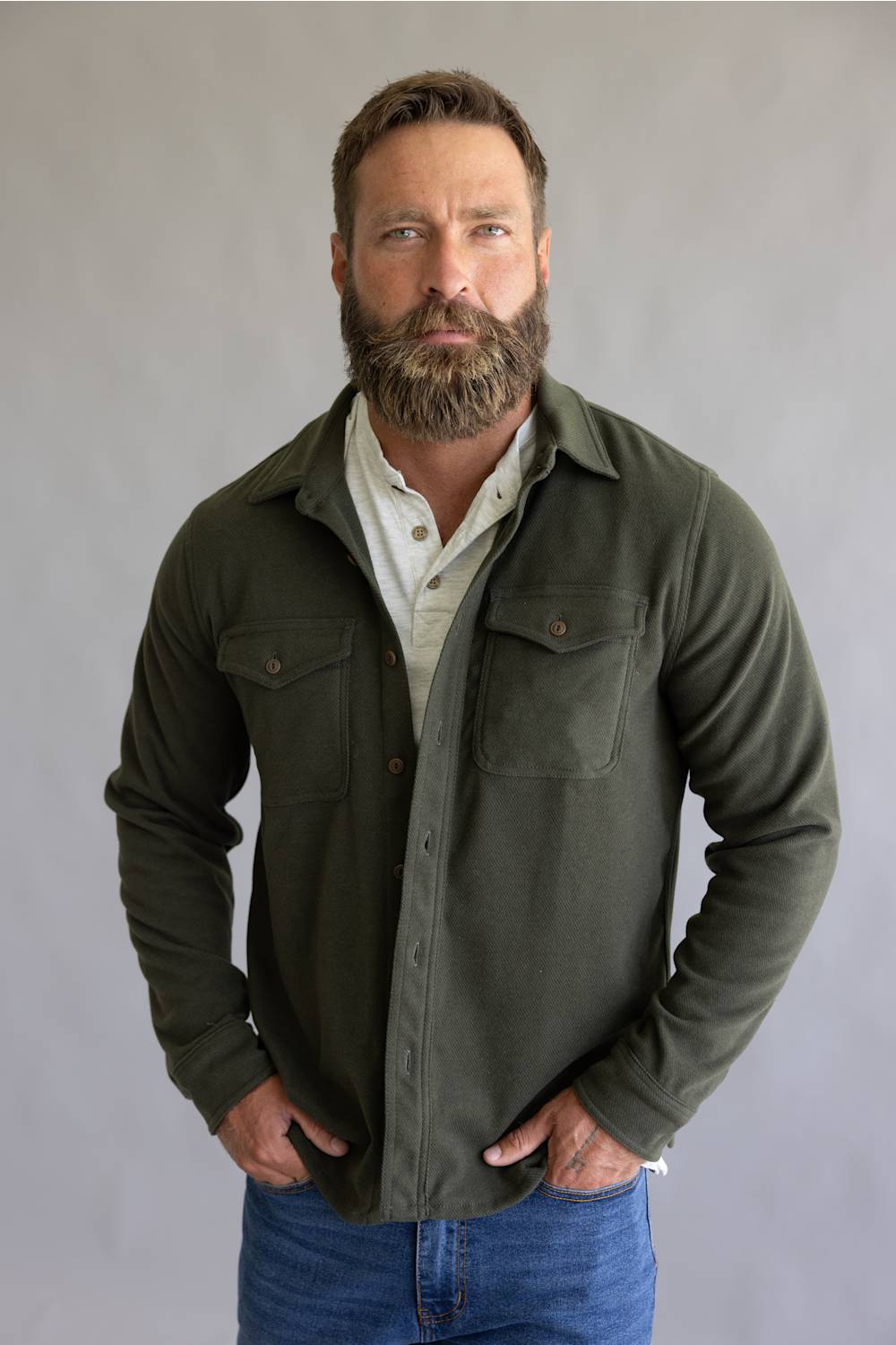 A man with a full beard wearing an olive green button-down shirt jacket over a cream button-down shirt and blue jeans, standing against a neutral gray studio background.