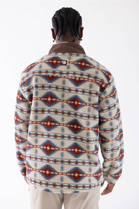 A man viewed from behind wearing a cream-colored sherpa fleece sweater with a red and blue geometric pattern, paired with beige pants, against a plain white background.