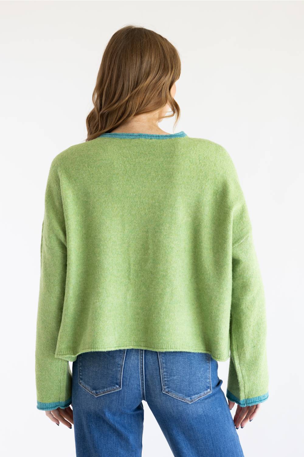 A woman viewed from behind wearing a light green cropped knit sweater with blue trim at the collar and cuffs, paired with blue jeans, against a plain white background.