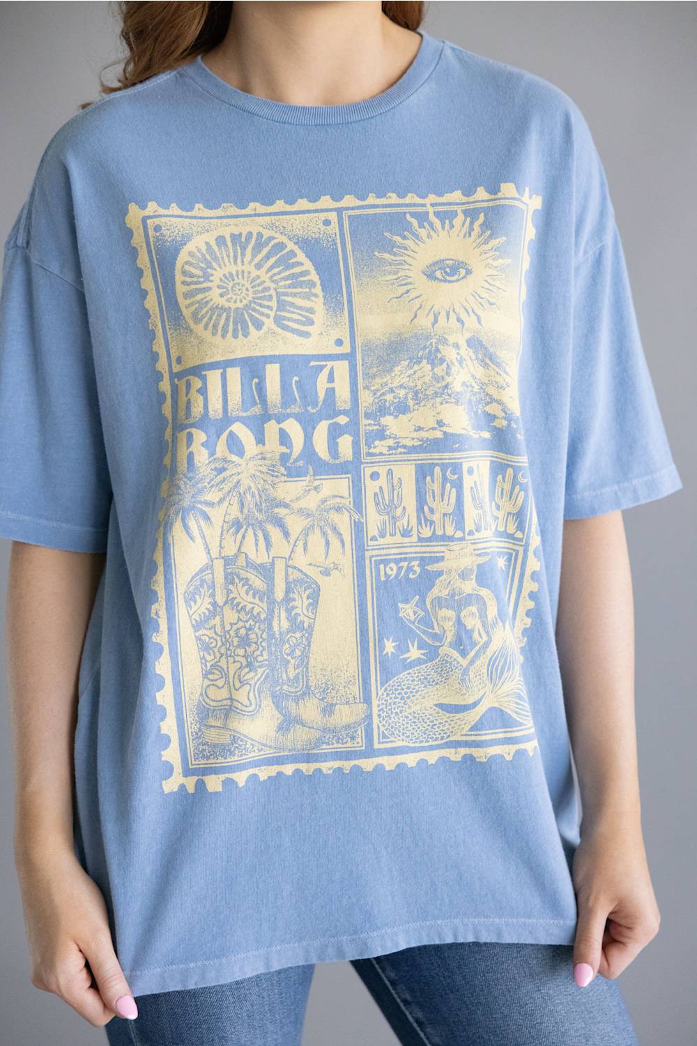 A person wearing an oversized light blue short-sleeve T-shirt with a large pale yellow Billabong graphic print on the front, paired with blue jeans against a plain gray studio background.