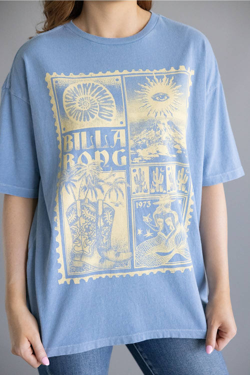 A person wearing an oversized light blue short-sleeve T-shirt with a large pale yellow Billabong graphic print on the front, paired with blue jeans against a plain gray studio background.
