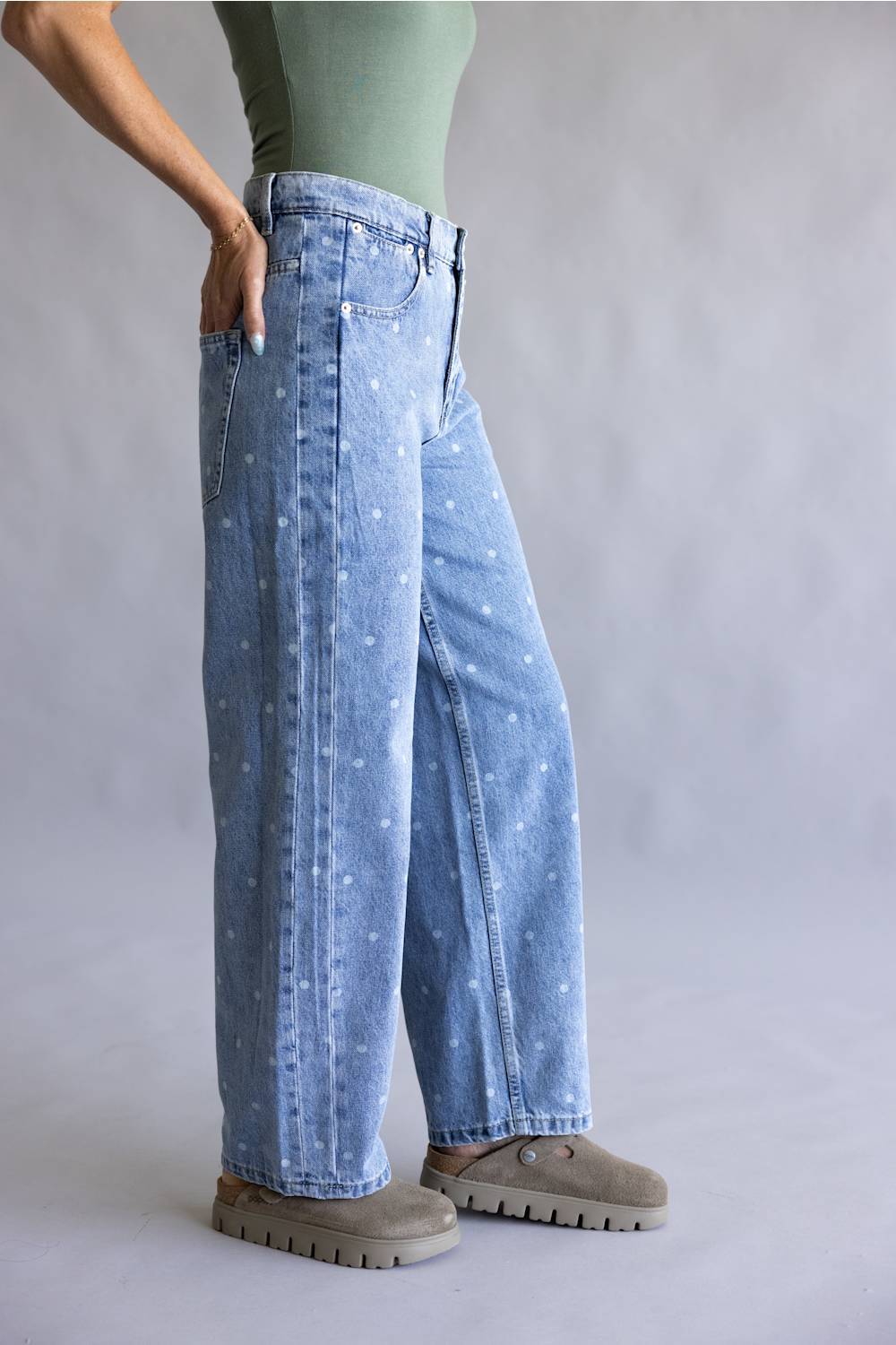 A person wearing light blue wide-leg denim jeans with a small white floral print, styled with a sage green top and brown suede boots against a neutral gray studio background.