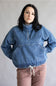 A woman wearing a blue denim half-zip pullover jacket with a high collar and front pockets, paired with dusty rose pants, standing against a plain light gray background.