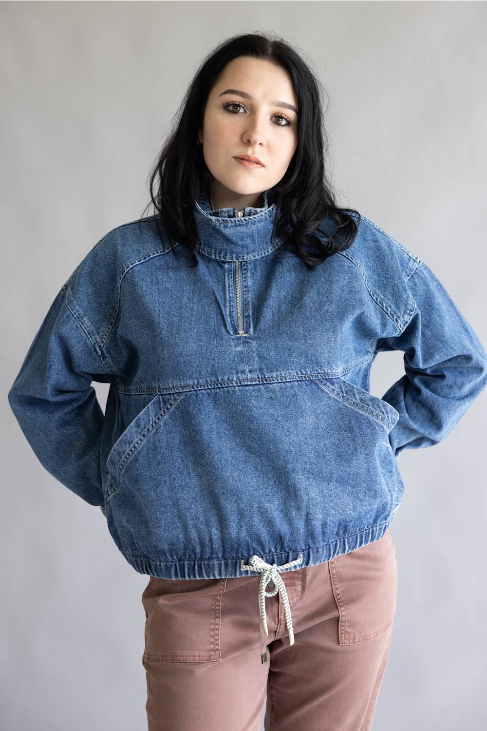 A woman wearing a blue denim half-zip pullover jacket with a high collar and front pockets, paired with dusty rose pants, standing against a plain light gray background.