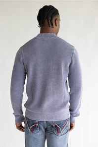 A man viewed from behind wearing a light purple ribbed knit sweater and blue jeans with decorative stitching on the back pockets, set against a plain white background.