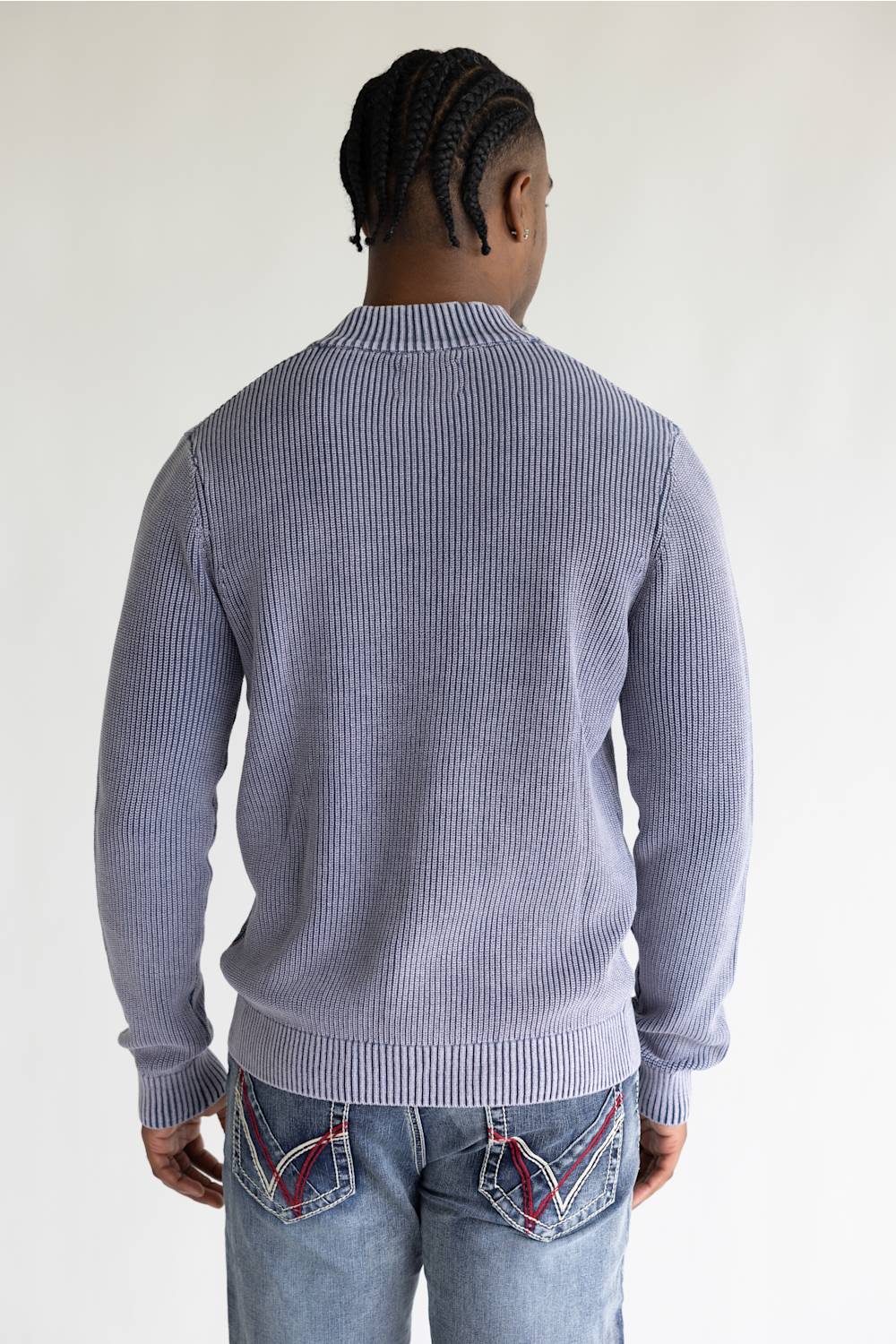 A man viewed from behind wearing a light purple ribbed knit sweater and blue jeans with decorative stitching on the back pockets, set against a plain white background.