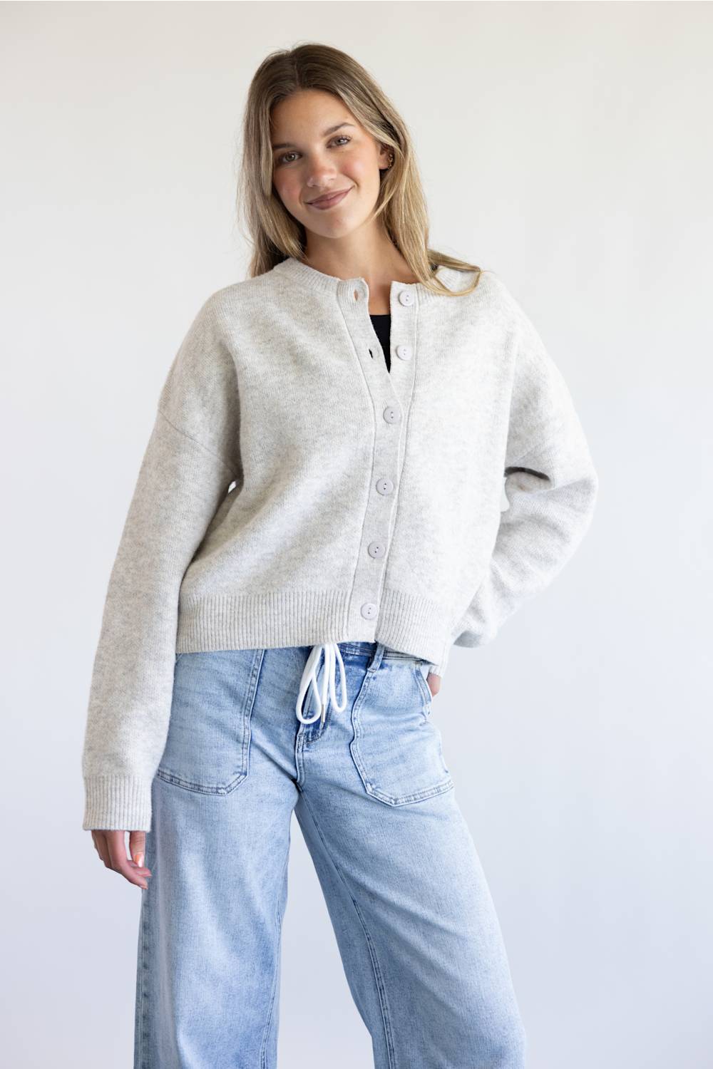 A woman modeling a light gray knit cardigan with a button-up front and drawstring detail, paired with light blue jeans against a plain white background.