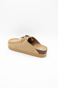 A single tan suede slip-on mule shoe with a small decorative black and white pom-pom detail on the side, set against a plain white background.