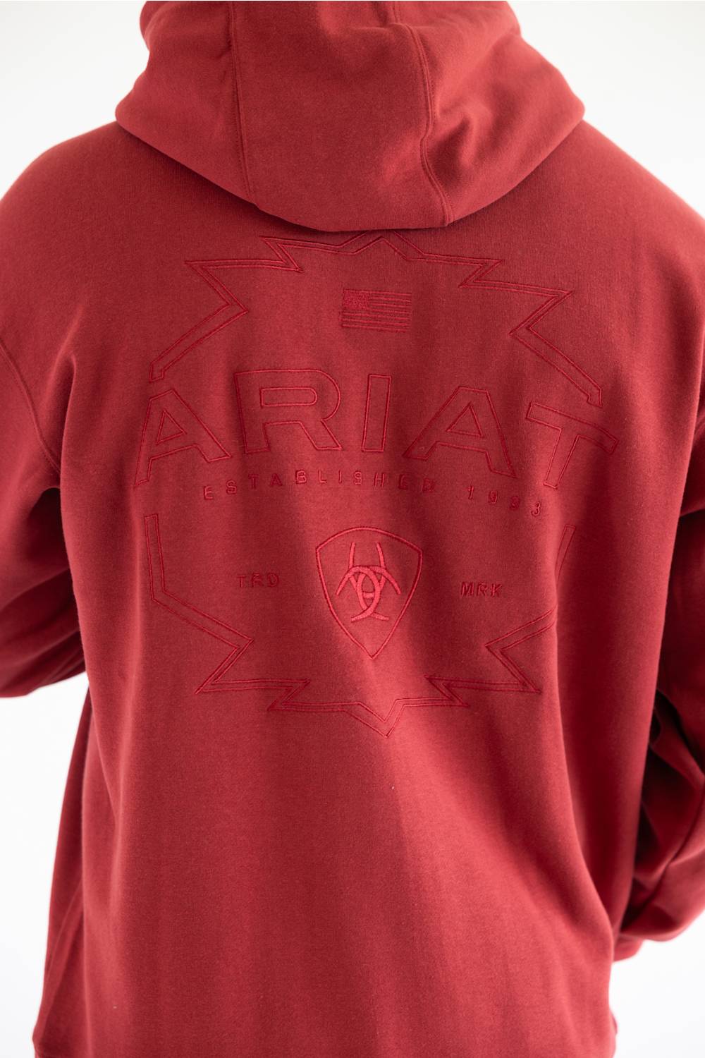 A close up of a man viewed from the back wearing a red pullover hoodie with a tonal embroidered logo across the back, paired with blue jeans, photographed against a plain white studio background.
