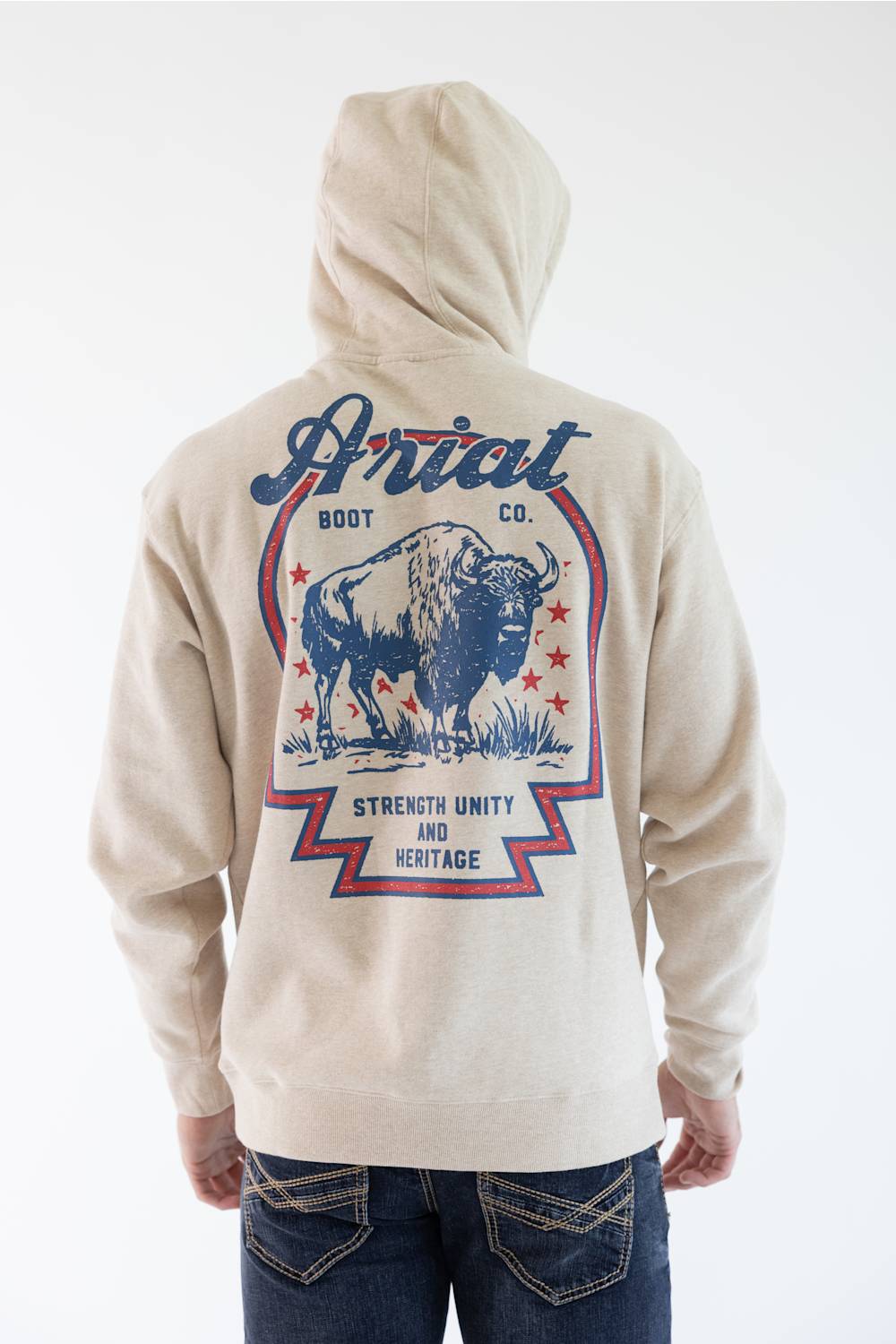 A person shown from the back wearing a light beige hoodie with a large blue and red graphic print featuring a bison and the text 'Ariat Boot Co.' and 'Strength Unity and Heritage,' paired with dark blue jeans against a plain white background.