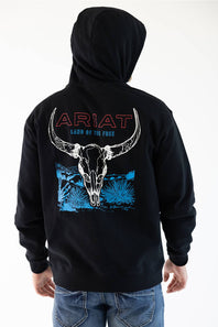 A person wearing a black hoodie from the back, featuring a large graphic print of a white bull skull over blue desert-style artwork, with 'ARIAT' and 'LAND OF THE FREE' text. The person is also wearing blue jeans against a plain white studio background.