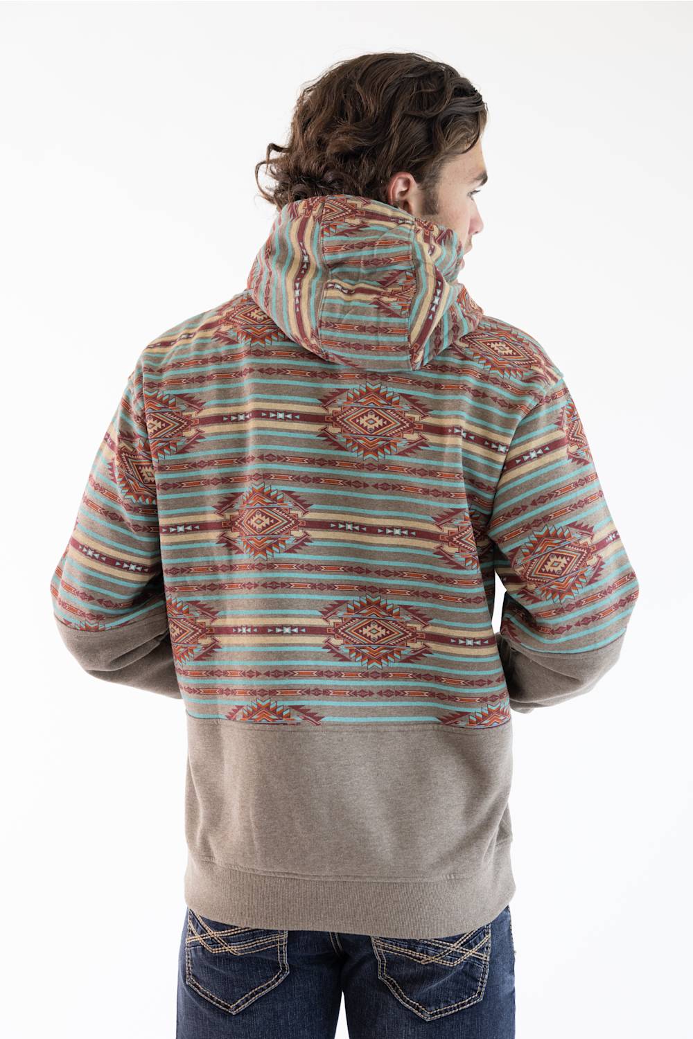 A man viewed from the back wearing a hooded sweatshirt with a multicolor geometric pattern on the upper body and sleeves, and a solid taupe lower body, paired with blue jeans against a white studio background.