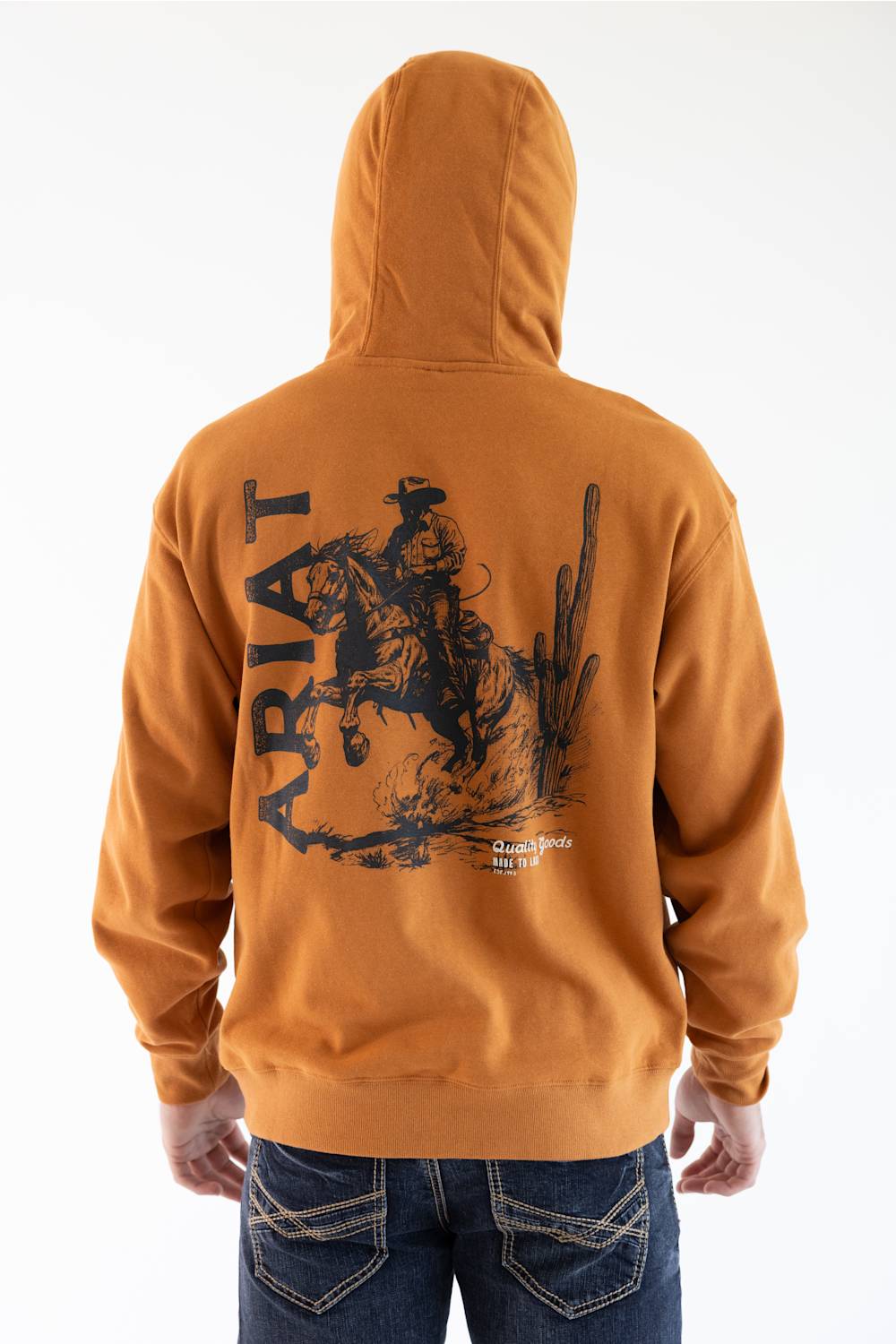 Close-up of the back of a burnt orange sweatshirt featuring a large black graphic print of a cowboy riding a bucking horse, with the word 'ARIAT' and desert illustrations.