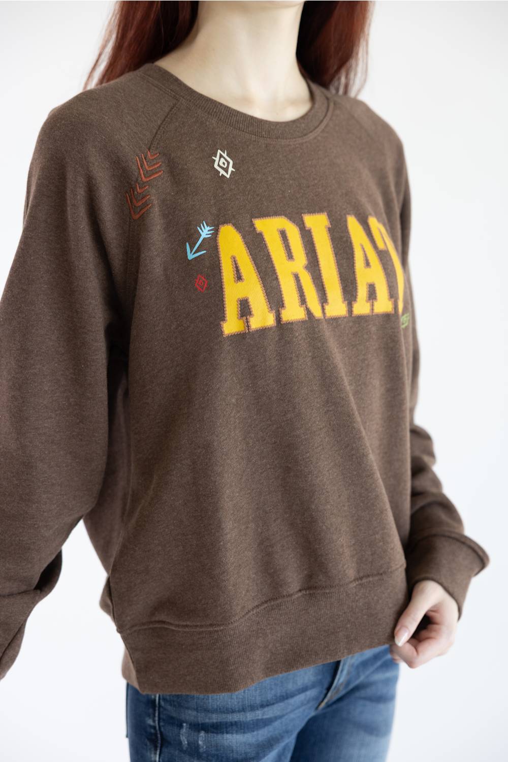 A woman with red hair wearing a brown crewneck sweatshirt with 'ARIAT' printed in yellow across the chest, paired with blue jeans, against a plain white background.