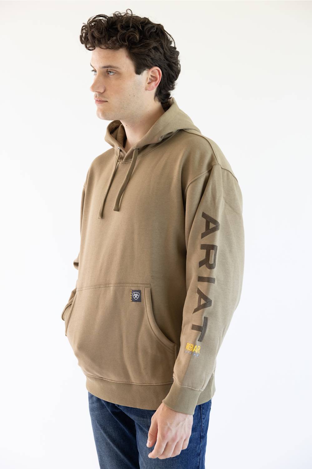 A man wearing a tan pullover hoodie with 'ARIAT' embroidered vertically in brown on the right sleeve and a small yellow logo patch near the cuff, paired with blue jeans against a plain white background.