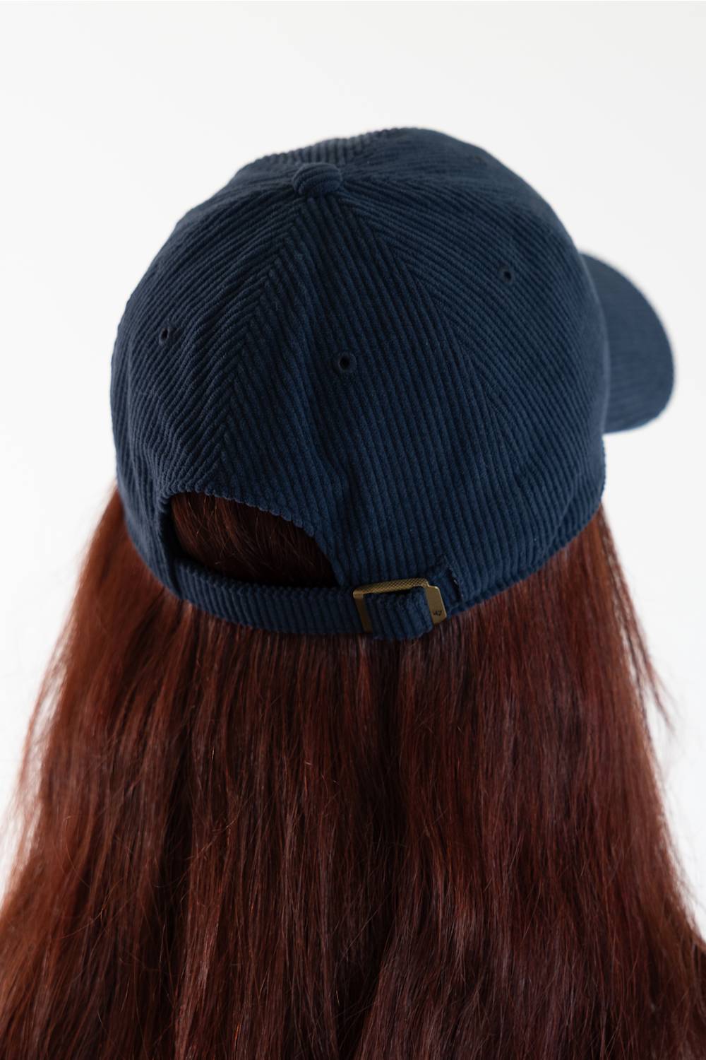 A dark navy blue corduroy baseball cap shown from the back, worn by a person with long, straight reddish-brown hair against a plain white background.