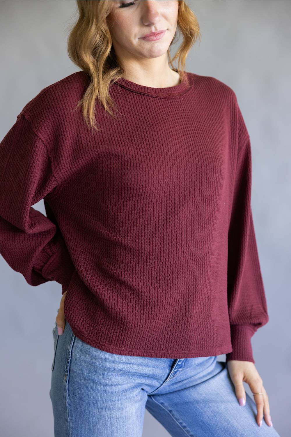 A woman wearing a textured, oversized crewneck sweater in burgundy with light wash blue jeans, standing against a neutral gray background.