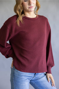 A woman wearing a textured, oversized crewneck sweater in burgundy with light wash blue jeans, standing against a neutral gray background.