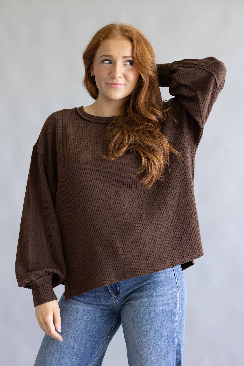 A woman with long red hair wearing a brown ribbed knit sweater with balloon sleeves and blue jeans, posing against a light gray studio background.