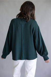 A woman viewed from the back wearing a dark forest green textured knit sweater with balloon sleeves and white pants, set against a plain gray studio background.